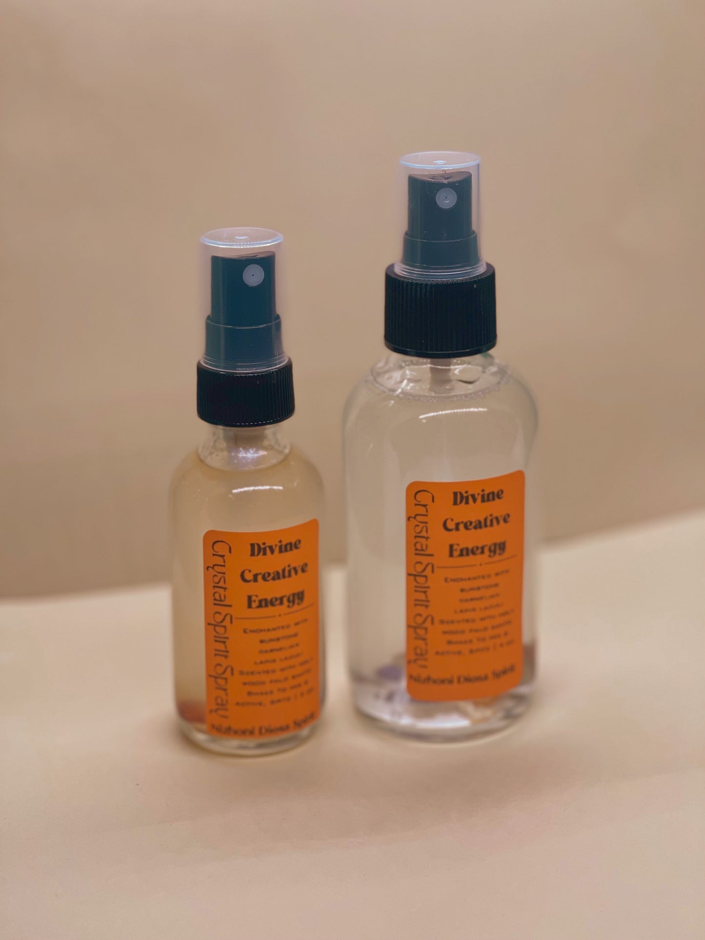 Two bottles of 'Divine Creative Energy' spray on a beige surface with a beige background.